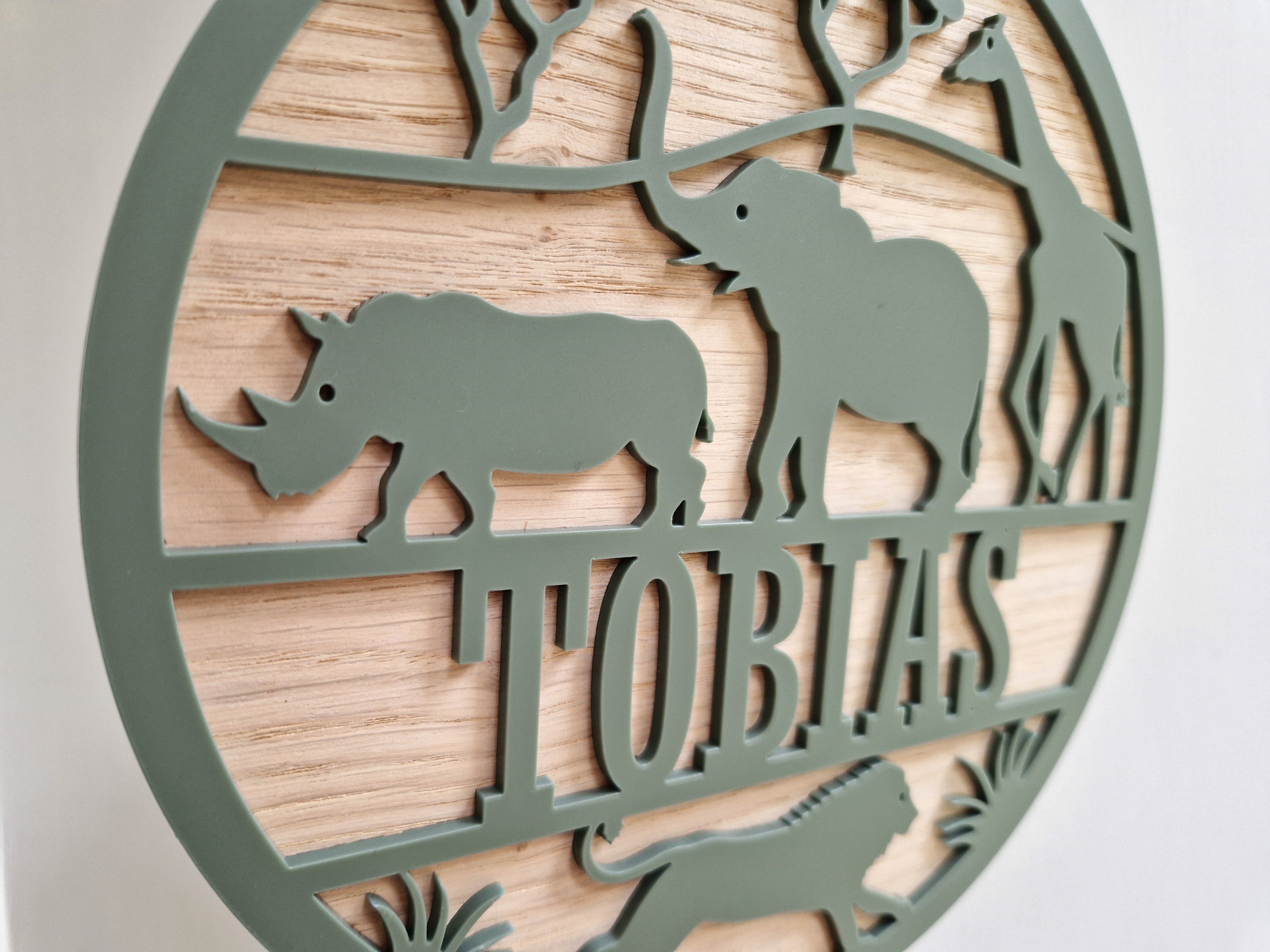 A circular metal sign with a wooden border, featuring silhouettes of a rhinoceros, an elephant, and a giraffe, along with the word "TOBIAS" written on it.