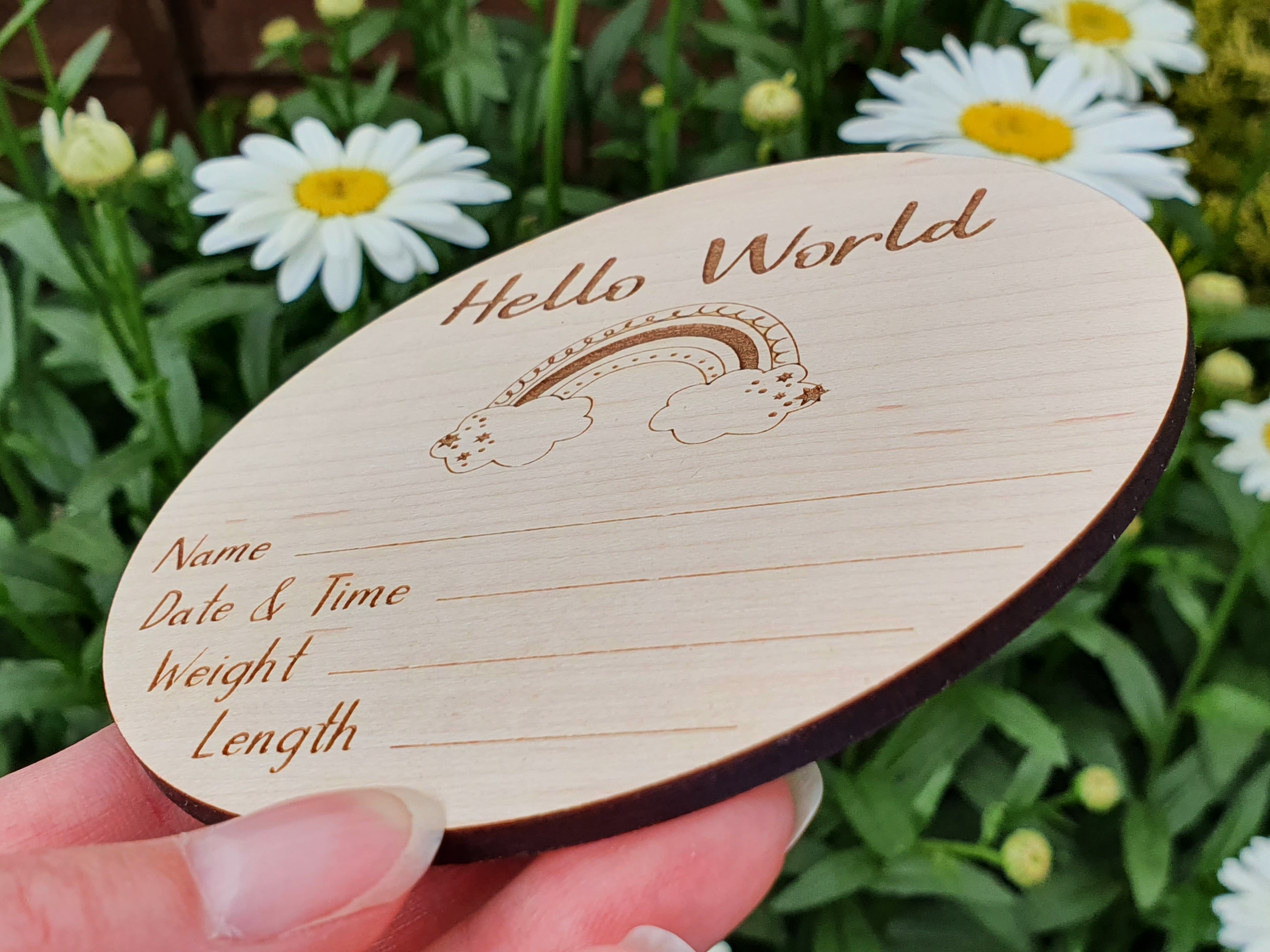 A wooden coaster or coaster-like object with the text "Hello World" and a drawing of a rainbow, with a person's hand holding it in front of a background of white flowers.