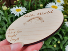 A wooden coaster or coaster-like object with the text "Hello World" and a drawing of a rainbow, with a person's hand holding it in front of a background of white flowers.