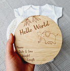 A wooden baby name card is being held up, featuring a cute elephant illustration and the text "Hello World" along with a blank space for the baby's name, date, time, weight, and length.