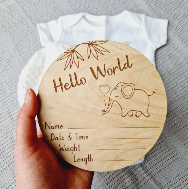 A wooden baby name card is being held up, featuring a cute elephant illustration and the text "Hello World" along with a blank space for the baby's name, date, time, weight, and length.