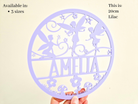 a hand holding a circular purple sign with a design featuring a fairy and flowers. The sign has the name "Amalia" written on it.