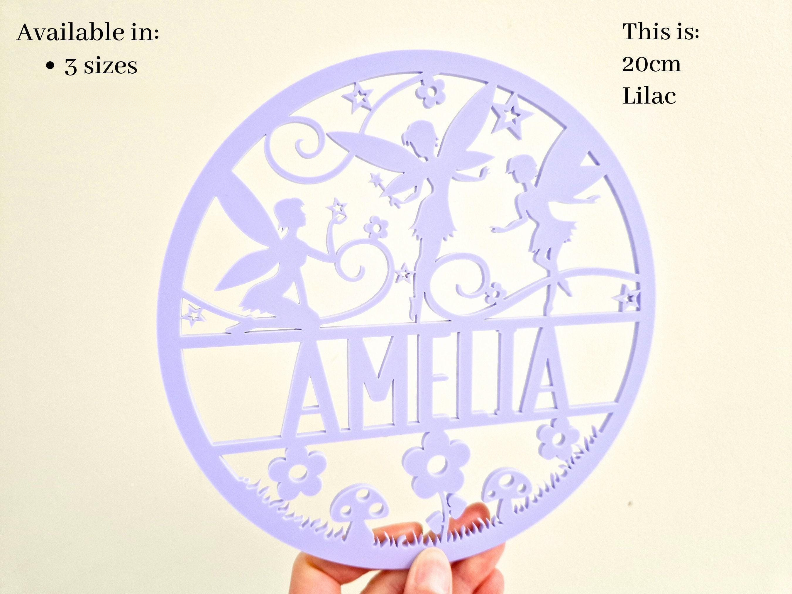a hand holding a circular purple sign with a design featuring a fairy and flowers. The sign has the name "Amalia" written on it.