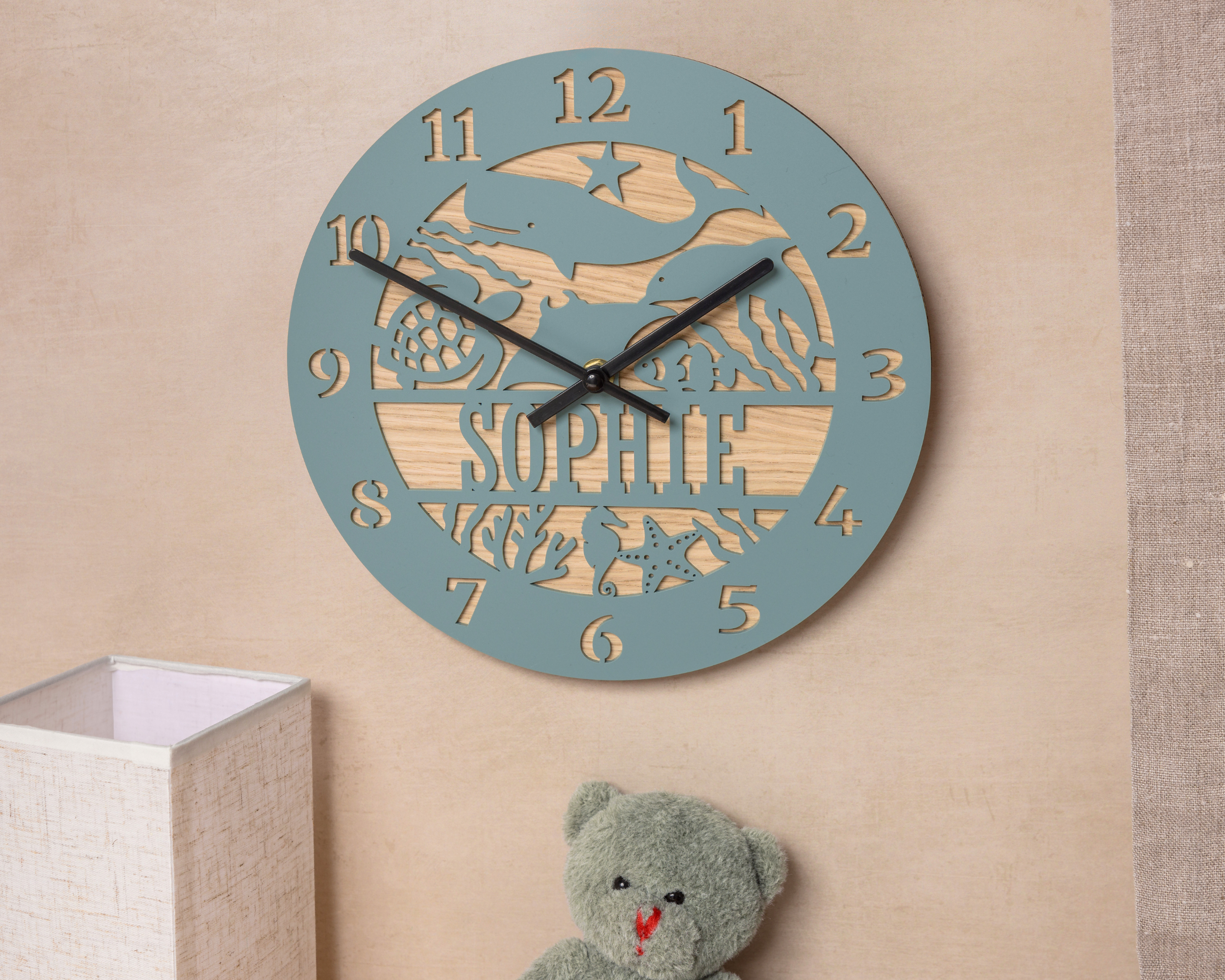 A wall clock with a blue and wood design, featuring the name "Sophie" on it, is mounted on a wall next to a stuffed teddy bear.