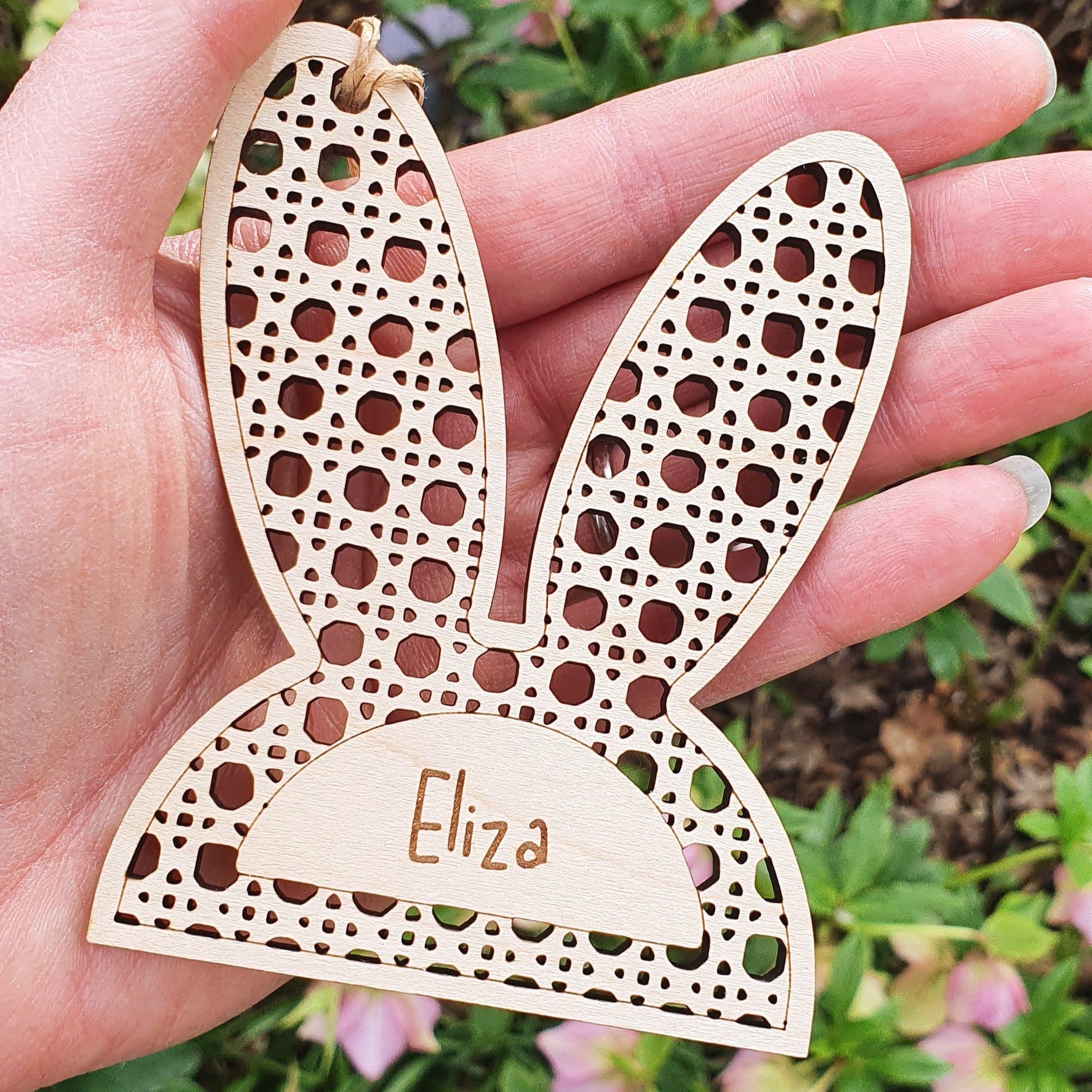 Wooden bunny ear-shaped ornament with 'Eliza' engraving held in a hand against a floral background.
